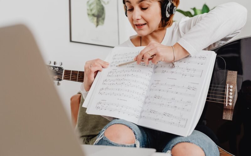 Pessoa tocando violão com um tablet aberto mostrando um curso online em um ambiente acolhedor e bem iluminado.