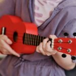 Mãos de um iniciante segurando um cavaquinho e tocando os primeiros acordes, com partituras ao fundo.