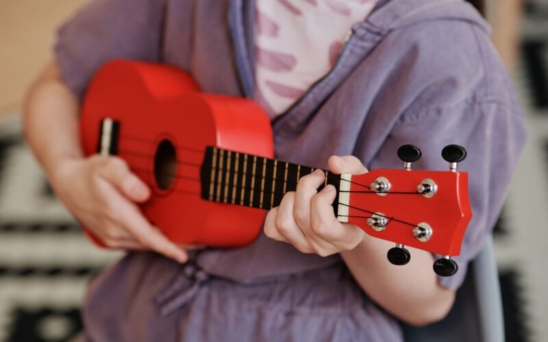Mãos de um iniciante segurando um cavaquinho e tocando os primeiros acordes, com partituras ao fundo.