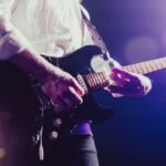 Homem tocando guitarra elétrica com fones de ouvido em frente ao computador estudando curso online.
