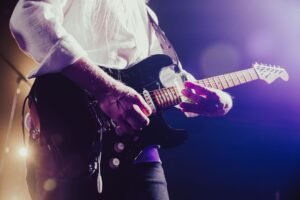 Homem tocando guitarra elétrica com fones de ouvido em frente ao computador estudando curso online.